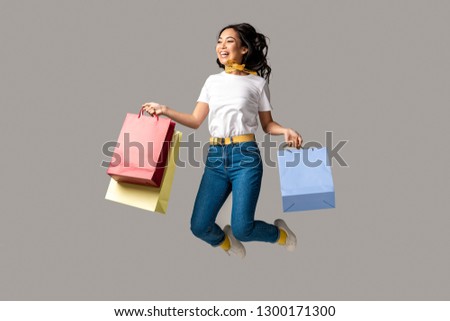 Laughing asian woman holding colorful shopping bags and happily jumping isolated on grey