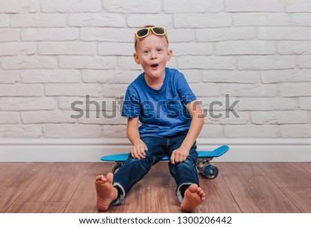 Portrait of a cheerful blond boy in a blue T-shirt, jeans and glasses on a blue skateboard