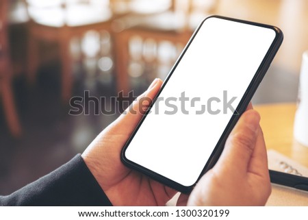 Mockup image of hands holding black mobile phone with blank white screen in cafe