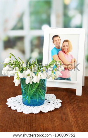 Bouquet of snowdrop flowers in glass vase, on wooden table, bright background