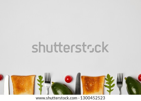 top view of toasts, avocados, cherry tomatoes, arugulas leaves, forks and knives on grey background