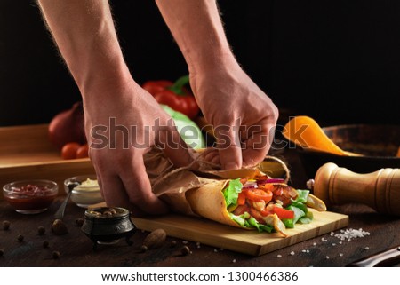 Shawarma sandwich in men hand on wooden rustic background. Gyro fresh roll of lavash or pita bread with grilled chicke, lettuce salad, cheese and vegetables. Tasty shawarma - middle eastern snack.