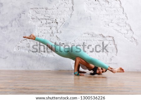 Side view - beautiful young brunette woman in gym clothes makes exercises for stretching  back and legs in an empty gym on white brick wall background. Space for text Royalty-Free Stock Photo #1300536226