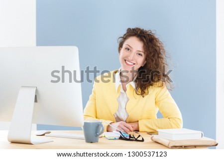 Attractive young female entrepreneur with red hair and spectacles in yellow jacked sitting at desk after hard working day, looking and smiling at the camera