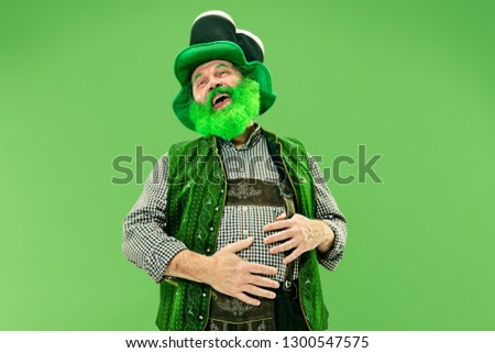 A smiling surprised happy senior man in a leprechaun hat with beard at green studio. He celebrates St. Patrick's Day. The celebration, festive, beer, holiday, alcohol, party, human emotions concept