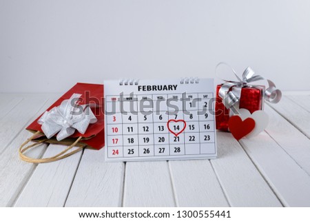 Calendar page with red hand written heart highlight on February 14 of Saint Valentines day with red gift bag, box and Heart shape on white wooden table