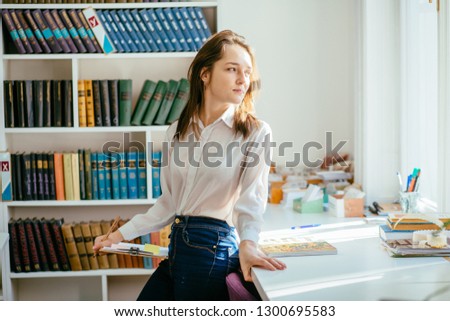 Beautiful thoughtful student girl sits with a notes on the windowsill with bookshelves on background.