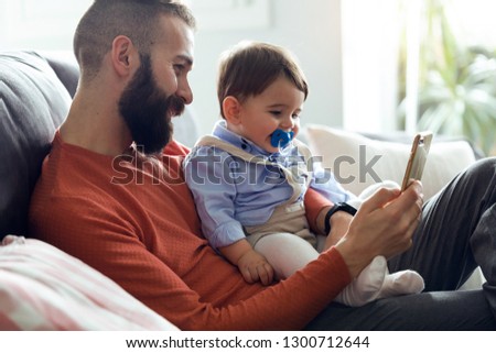 Shot of handsome young father with his baby looking cartoons on mobile phone on sofa at home.
