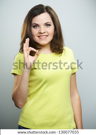 Happy young woman in yellow shirt and jeans. Woman showing her right hand okay. On a gray background