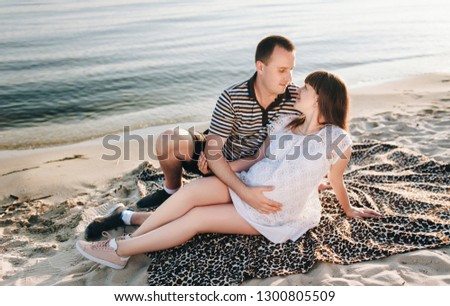 A pregnant red-haired girl in a white dress and a young beloved boy hugging, sitting on a plaid, on the beach. Waiting for a child. Pregnancy girl. Tender picture on the beach with sand.