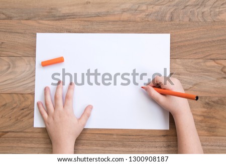 Child's hands with orange felt-tip on the wooden table with a piece of paper. Top view.