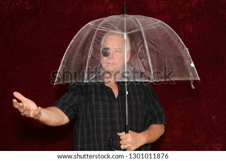 Man with an Eye Patch and an Umbrella. A man poses in a photo booth with a Burgundy Red Velvet Background with a clear umbrella. Room for text. 