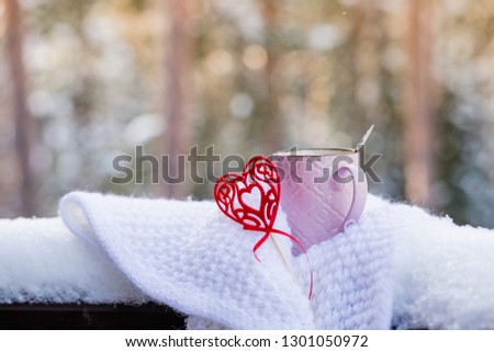 Cup with red heart and a hot drink wrapped in a scarf in the snow. Concept for Valentine's day.Romantic Morning breakfast for Valentines day on snowy forest background.Hello February. vintage red