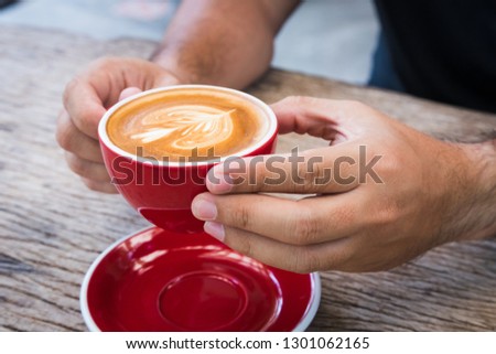 man hand hold red cup of coffee with milk making for flower picture on plate and spoon then lift it up with two hands on bark wooden table