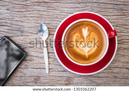 coffee with milk making for flower picture in red cup on plate and spoon put on bark wooden table, with black smartphone