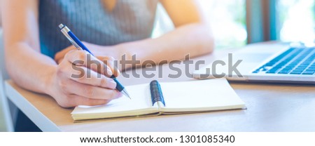 Woman hand is writing on notepad with pen in office.On her desk there is a laptop.Web banner.