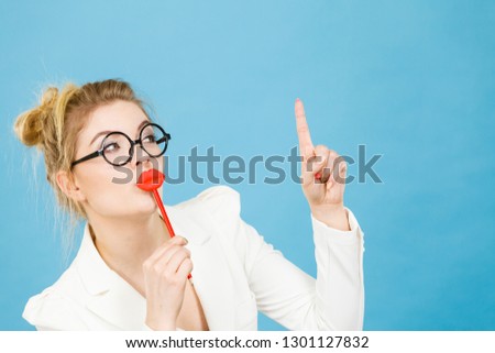 Lovely sweet business woman elegant clothing nerdy glasses holding red fake lips on stick having fun, pointing up with finger at copy space, on blue. Carnival funny accessories concept.