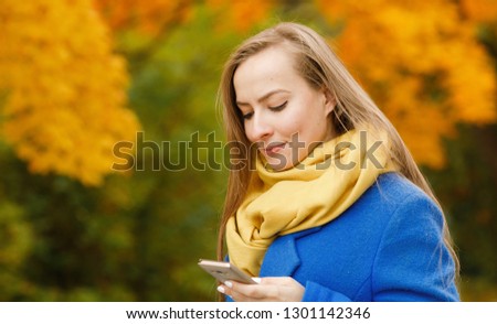 Emotional portrait of a positive and happy beautiful girl looking with a smile at the smartphone against the background of blurred yellow foliage on a walk in the autumn park. Lifestyle. Autumn mood