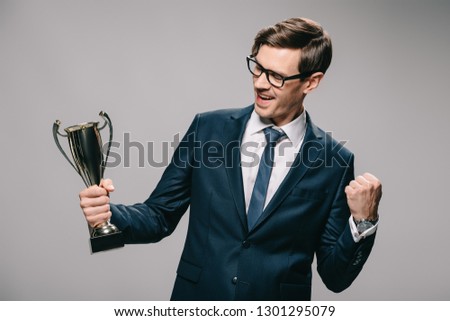 cheerful businessman in glasses celebrating victory and holding trophy in hand on grey background