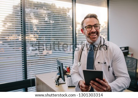 Smiling mature male doctor with digital tablet in his office. Friendly medical professional with tablet computer in clinic. Royalty-Free Stock Photo #1301303314