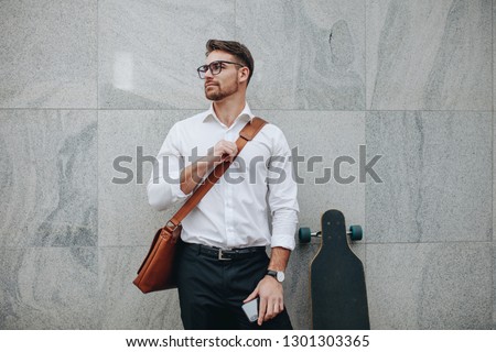 Businessman wearing an office bag standing against a wall holding mobile phone. Formally dressed man standing with a long skateboard looking away.