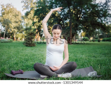 Young pregnant concentrated woman stretching outside in park. She hold one hand up and another on belly. Model look down. She sit in lotus pose.