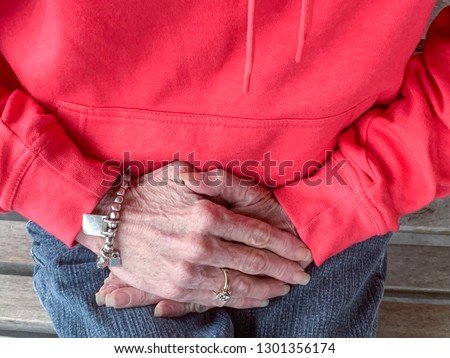 A Caucasian woman in red pull-over shirt and blue jeans sitting on a weather-beaten bench prominently featuring her veiny hands.