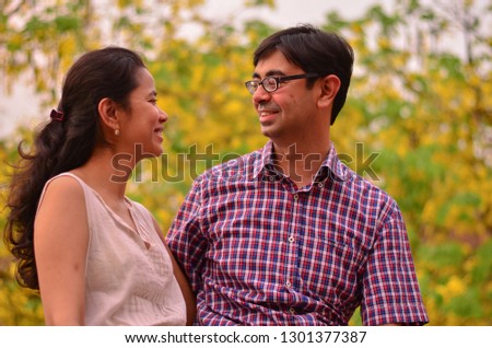 Young Indian couple looking at each other and smiling standing against yellow amaltas (golden shower) tree leaves during summer autumn weather in Delhi, India
