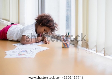 Mixed race girl, tanned skin, Curly short hair lying down, drawing and painting with concentration and intended at the window in the classroom in the morning. In Bangkok, Thailand. Education Concept