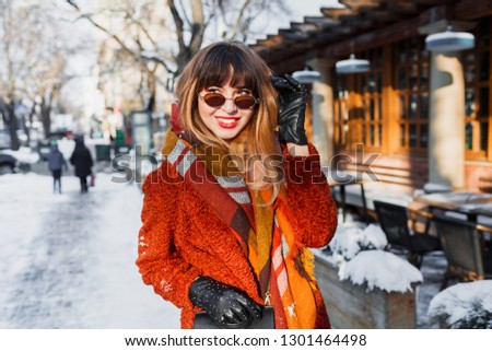 Fascinating happy woman in  retro glasses posing outdoor in old European city. Standing near cafe. Wearing brown  coat, wool scarf and leather gloves.