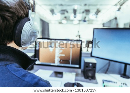 Young man with headphones is sitting in the recording studio, buttons, studio, screens and equipment in the blurry background