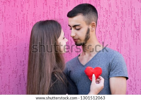 Waist-up portrait of sweet couple standing together on pink background. Charming dark-haired girlfriend holding soft red heart. Love and relationship concept