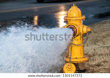 Close-up of yellow fire hydrant gushing water across a street with wet highway and tire from passing car behind Royalty-Free Stock Photo #1301547373