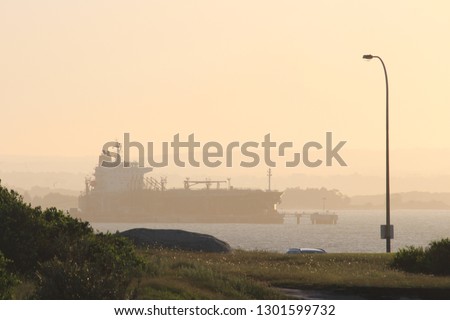 Landscape looking out over the sea to silhouette of land and a docked ship. The ship is unloading its cargo of petrol. A soft orange sky sunset grass and rocks in the foreground. La Perouse, Sydney