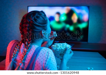 A young girl watching movies and eating popcorn with a bowl on the background of the TV. The color bright lighting, blue and red. Relax, rest at home when watching TV, film. Background for design