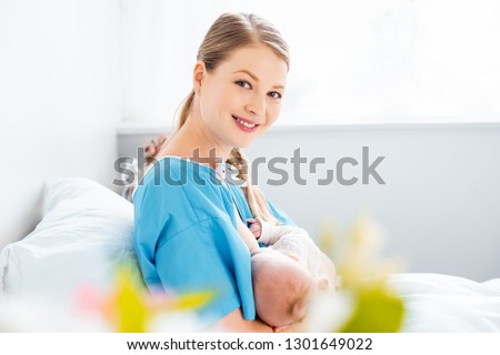selective focus of happy young mother breastfeeding newborn baby and smiling at camera in hospital room