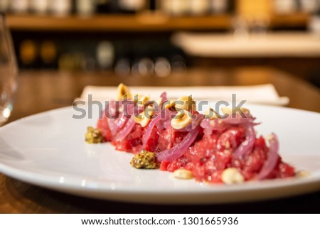 A wooden table with local beef tartare, pickled onions and hazelnuts on a white plate, red wine and some bread. The picture is took from above.