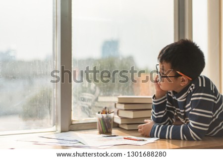 The boy sitting by the window is using his thoughts and gaze to look outside, doing his homework intently, writing and painting on the table in an art class. 
