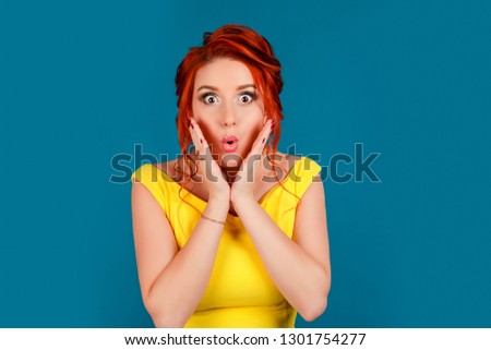 Surprised pretty woman with bright make up. Half body portrait of Caucasian person in yellow dress, redhead hair up isolated on blue studio background. Expressive girl, human emotion, face expression