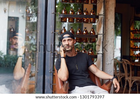 Indoor shot of handsome trendy looking college student in stylish clothes and accessories having serious phone conversation while drinking cappuccino at coffee house, sitting by window alone.