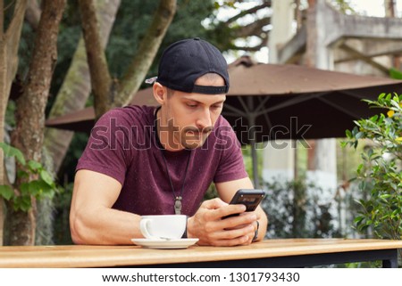 Handsome man in casual sporty clothes holding phone, writing notes in mobile application, biting lower lip while trying to concentrate on work sitting at cafe. University student busy with preparation
