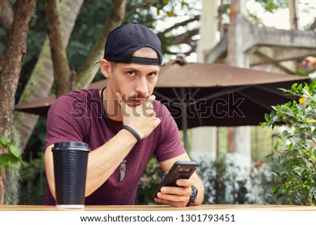 Thoughtful Caucasian handsome man touching chin, focused on finding new approaches and solutions in business while chatting at modern cafe, sitting at table outdoors with mug and mobile phone in hand.