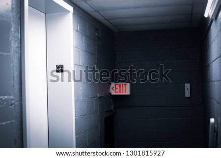 A cold, dark, haunted hallway in a building with an elevator, a door and an exit sign. 