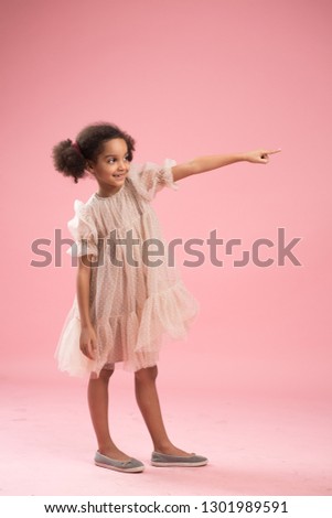 Positive smiling ethnic woman has Afro haircut, dressed in fashionable dress, points with thumb at blank copy space, poses against pink background. Ethnic black lady advertises new item indoor