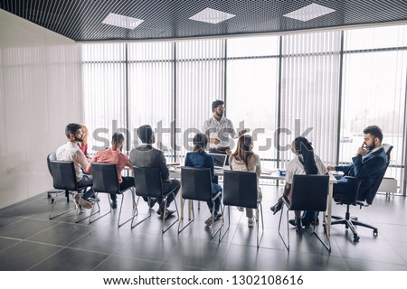 Achieving best results after annual refresher course. Indian young business mentor conducting a business training while standing in front of people sitting in row at meeting room. Royalty-Free Stock Photo #1302108616