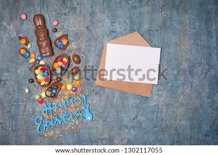 Top view on mockup of blank white card in kraft envelope with text of happy easter and chocolate eggs and bunny on blue concrete background