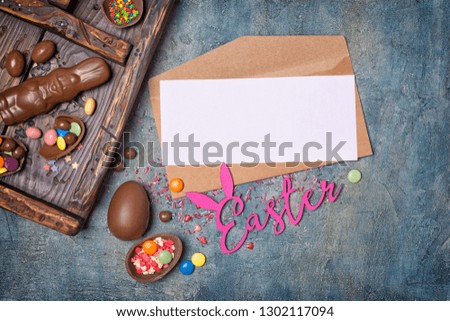 Top view on mockup of blank white card in kraft envelope with chocolate eggs and bunny in vintage wooden box on blue concrete background