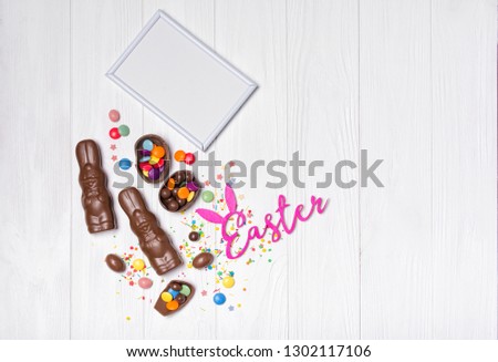 Top view on mockup of blank white frame with text of easter and chocolate traditional eggs, bunny and color dragee on white wooden background with copy space