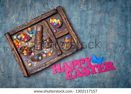 Top view on chocolate traditional eggs, bunny and sugar sprinkles or confetti in vintage wooden box with bright text of happy easter on blue concrete background