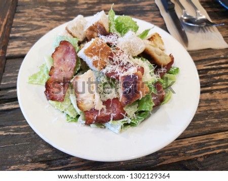 a white plate of caesar salad on cracked wooden table. grilled bacon, cos lettuce, scorched croutons and parmesan cheese on dish. In the top corner of picture there was knife, spoon and folk on tissue